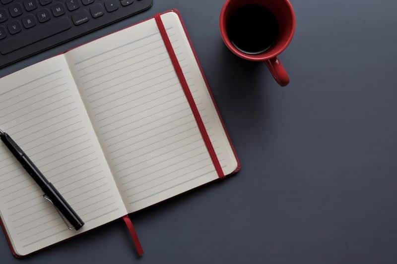 Agenda, parte de un teclado y un café en una taza roja en un escritorio gris oscuro